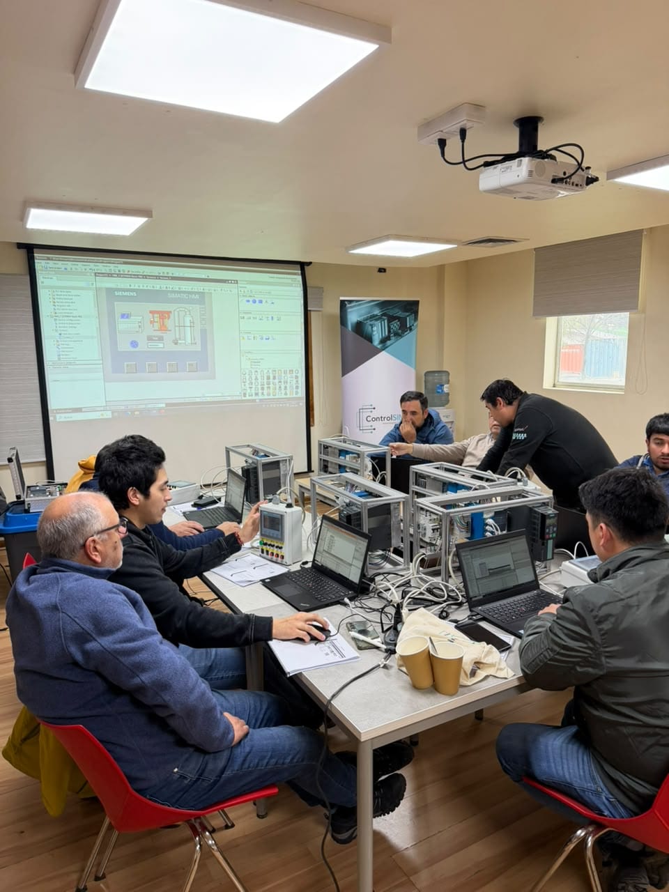 Participantes programando en TIA Portal en sus estaciones de entrenamiento, con el diseño de una pantalla SIMATIC HMI proyectado y el pendón de ControlSIM al fondo